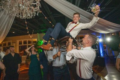Wedding guests lift the groom in a chair during a celebratory tradition at The White Room venue in historic downtown St. Augustine. The groom, wearing a white shirt with suspenders and bow tie, raises a glass while being hoisted up by groomsmen in white shirts. Wedding guests watch and cheer in the elegantly decorated reception space with crystal chandeliers and draped fabric.