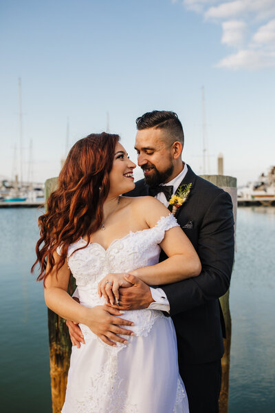 Newly married couple embraces each other while in a tux and gown on a pier
