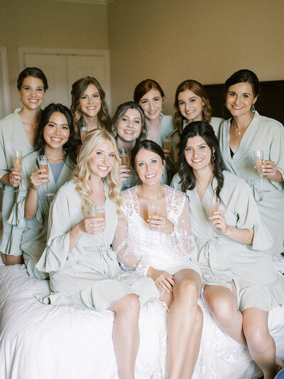 bride and her bridesmaids all sitting on a bed together with champagne in hand after finishing services with Indianapolis makeup artists and hairstylists, Something Blue Stylists