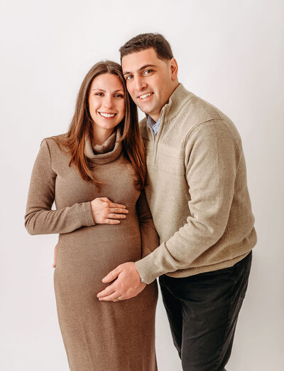 Studio maternity portrait by Stamford CT photographer capturing expecting couple in matching neutral sweaters with hands gently placed on baby bump