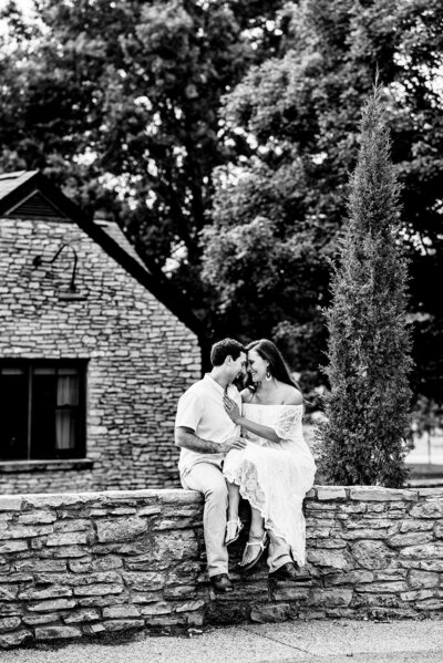 Black and White photo of couple sitting on rock wall, nuzzled together forehead to forehead