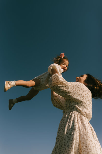 A mother playfully swinging around her daughter high in the air with the deep blue sky as their background. 