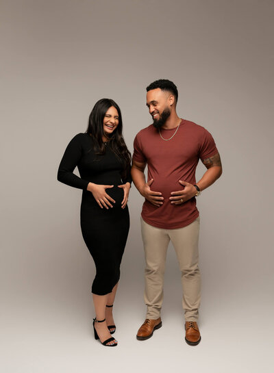 pregnant mom holding her belly and laughing in black dress while husband holds his belly