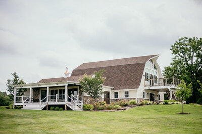 A large, white barn-style building with a brown roof sits on a lush green lawn. It features a wraparound wooden deck, expansive windows, and is embraced by trees and shrubs under a cloudy sky—a picturesque setting reminiscent of the tranquil landscapes at Halcyon Watson.