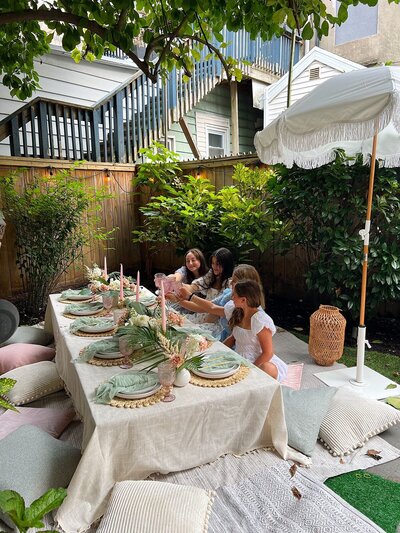 Backyard celebration with a styled picnic table, pastel cushions, candles, and floral centerpieces, designed by Yours Truly, a wedding planner and designer in New Jersey.