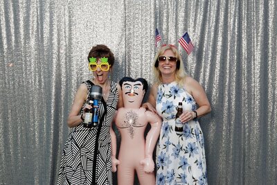 Women in photobooth with silversparkly background, holding a blow up doll and silly props