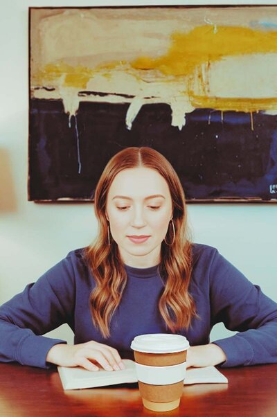 Stacey Xanthe in college, sitting at a desk reading a book with a coffee cup in front, wearing a navy sweatshirt with a Rothko-inspired painting in the background.