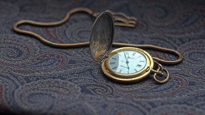 A vintage gold pocket watch with its cover slightly open, revealing Roman numerals, placed on a richly textured paisley fabric.