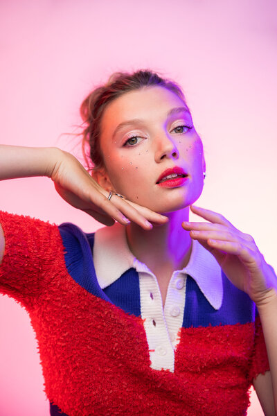 Portrait of a woman in a red, white and blue collared shirt with her hair up in a messy bun and a soft pink hue in the background.
