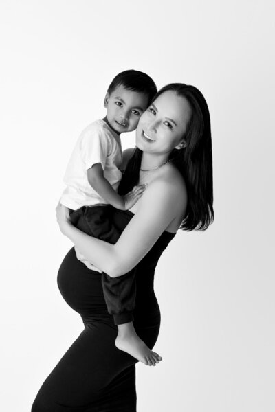 A pregnant woman smiles while holding her young child in her arms against a white background. She is dressed in a form-fitting black dress that highlights her baby bump, while the child wears a white T-shirt and black pants. The woman gazes lovingly at the camera, and the child rests comfortably on her hip, looking directly at the camera. The black-and-white composition emphasizes their joyful connection.