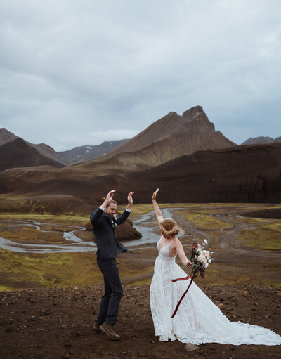 groom and bride are standing next to each other looking out into the distance at an alpine lake in breckenridge. the bride is holding her flowers and she has her hand on the groom's shoulder. there are mountains with snow in the background.