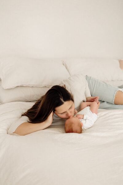 A woman in a cream color sweater poses in her living room in Colchester, Vermont. She sits smiling while holding her sleeping newborn baby. A perfect setting for capturing cozy moments by a New England Portrait Photographer.