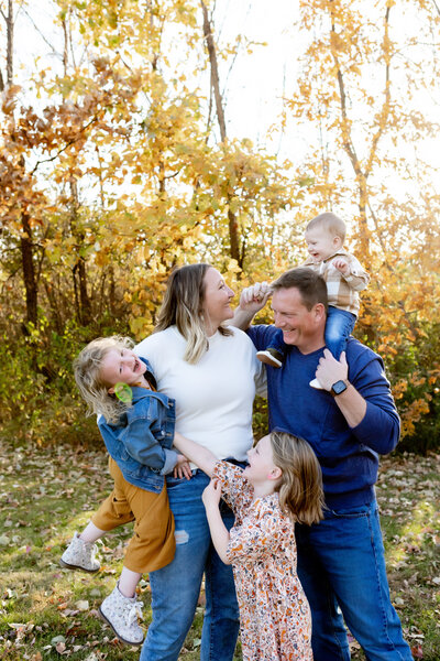 A joyful family of five shares a playful moment outdoors in the fall, surrounded by golden autumn leaves. The mother holds one child in her arms while laughing with her partner, who carries their youngest on his shoulders. Another child reaches up toward them, smiling. The warm sunlight filters through the trees, enhancing the lively, affectionate atmosphere.
