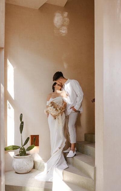 Bride and groom share an intimate kiss on the staircase of a private villa in Tulum, with minimal desert décor and potted cactus.