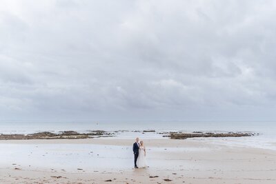A bride and groom standing on the sand at the beach with rocks and ocean in the background