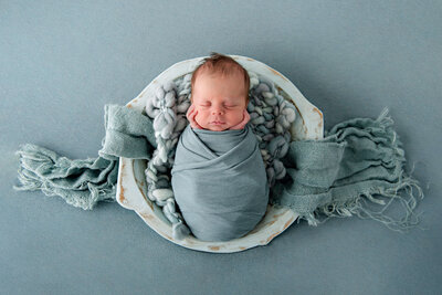 baby boy wrapped in blue laying in a blue bowl on a blue background for his newborn photos in Burlington, Ontario.