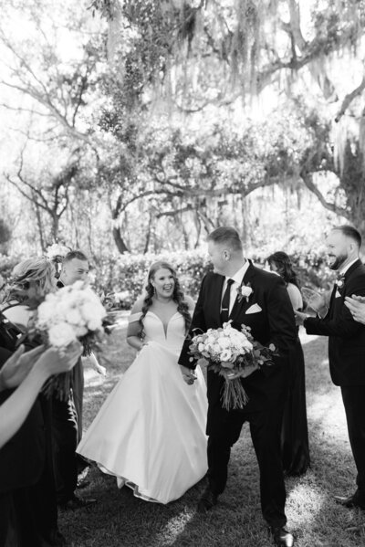Blonde Bride walks back down the aisle with her groom in a navy suit. The weather is cool and there is a large tan leaf tree decorated with white drapery and florals in the backyard.