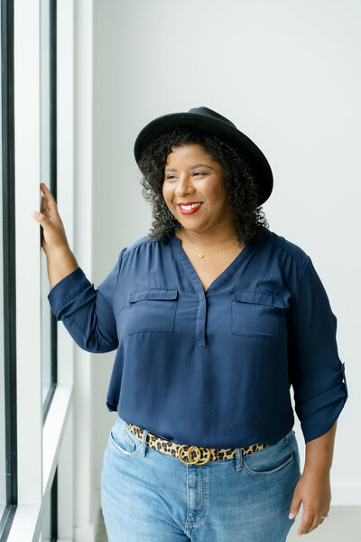 A bright and inviting portrait of Mica McCook, standing by a window and smiling while wearing a blue top and a black hat. Captured by a fellow photographer, this image highlights Mica’s approachable style and her love for natural light.