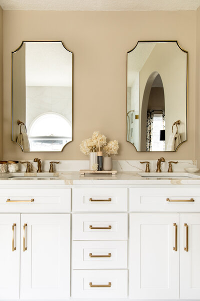 Large white double sink vanity with gold mirrors, hardware and plumbing fixtures featuring a neutral beige wall paint.