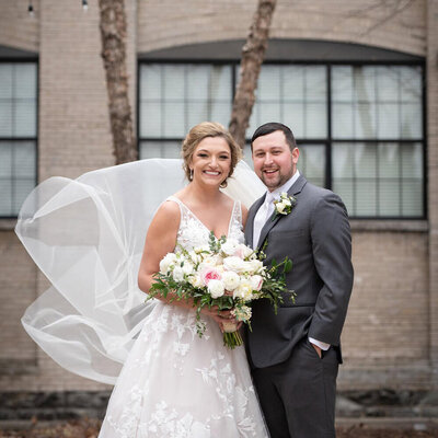 bride with flowing veil and groom stand in front of tree and building