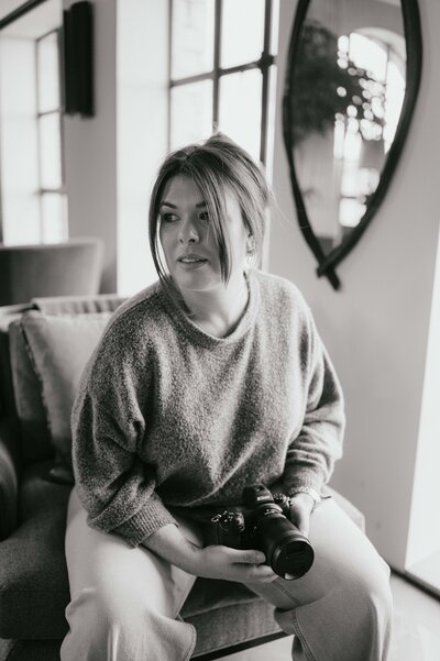 Sarah Hurja sitting on a sofa, holding a nikon camera and looking away pensive.
