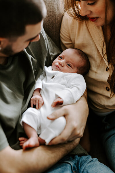 New parents hold newborn baby boy and look down on him with smiles during lifestyle newborn session in Nashua, new Hampshire