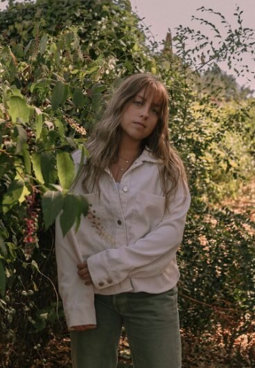A person standing up against bushes with one arm across their body