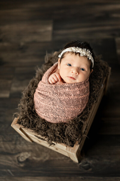 Newborn baby girl bundled in a pink wrap, sitting inside a wooden box, on a dark wooden floor.