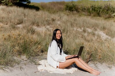 Girl smiling at the camera whilst working on her laptop at the beach