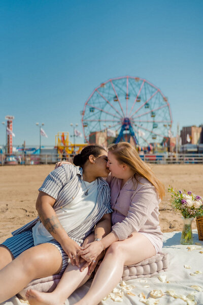 couple sitting on a blanket on the beach kissing with a ferris wheel behind them