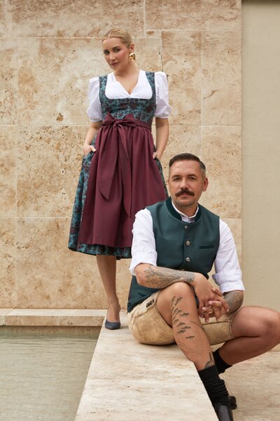 Paar in moderner Designer-Tracht von Monaco Spatzl: Frau im floralem blau-dunkelrotem Dirndl und Bordeauxroter Seidenschürze, Mann in blauer Weste von Monaco Spatzl, Hirsch-Lederhose der Marke Schweigerts  – hochwertige Trachtenmode aus München.