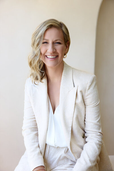 woman sitting on a desk in a light suit looking into the camera smiling in the studio