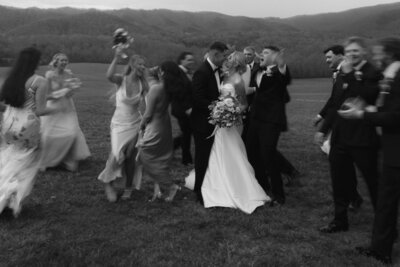 A black and white photo of a groom kissing a bride on the cheek while wedding guests in the background cheer and clap. The setting appears to be outdoors with rolling hills in the distance.
