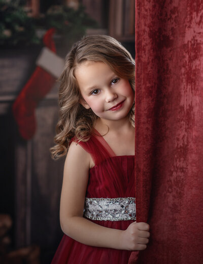 A young girl stands behind a red curtain looking at her Iowa mini photographer. 
