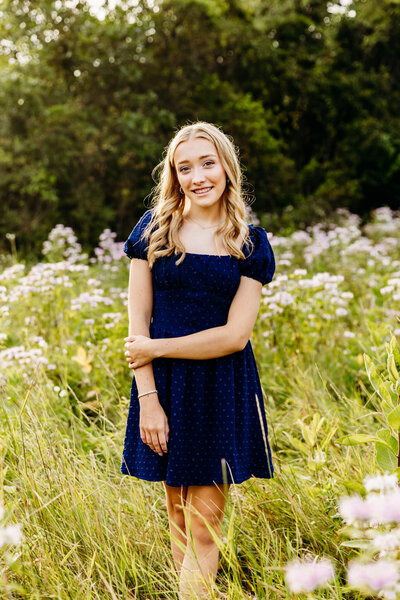 stunning blonde senior girl in a blue dress standing in a bright green field filled with purple bee balm captured by Ashley Kalbus Photography