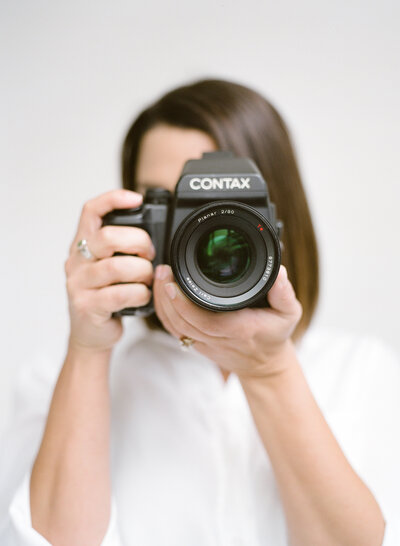 Photographer holding camera up in front of face on white backdrop