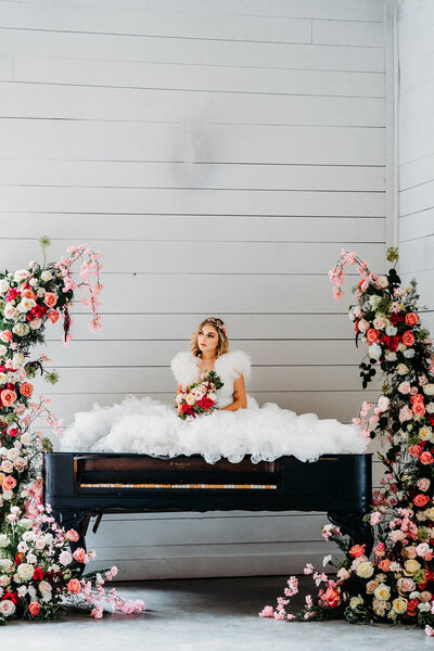 a bride sitting on a piano with 2 floral installations on both sides of the piano