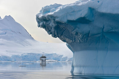 A large iceberg with a rugged surface floats in calm polar waters, with snowy mountains and a small ship visible in the distance.