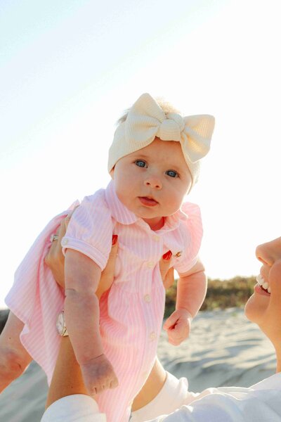 A mother playfully holds her baby girl in a pink dress above her head at the beach, enjoying a sunny day together.