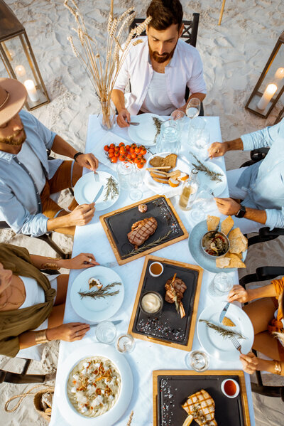 Five people seated at a beachside table adorned with blue and white linens, enjoying a meal. The table features grilled meat on slate plates, a salad, bread, tomatoes, and decorative plants. Beach sand and candles surround the area.