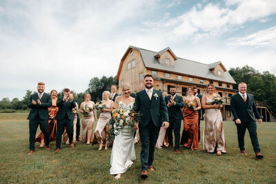 Bride and groom walking with their wedding party in front of the rustic barn venue at Bear Ridge Destination on a cloudy summer day.

