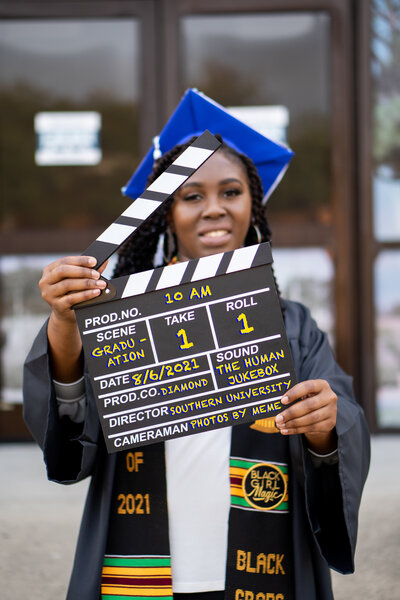 Graduate hold directors clapperboard while wearing graduation cap and gown