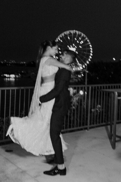 Groom lifting his bride during nighttime wedding portraits with a glowing ferris wheel blurred in the background.
