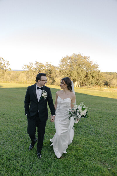 A full-body shot of a bride and groom walking hand-in-hand across a grassy lawn on a sunny day. The groom wears a black tuxedo with a bow tie and glasses, while the bride wears a white wedding gown and holds a bouquet of white roses.