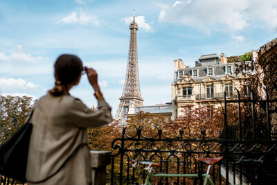 A person stands in the foreground, contemplating the Eiffel Tower in Paris. The scene captures classic Parisian architecture, trees with autumn foliage, and a bike against a black wrought-iron fence under a partly cloudy sky—a perfect snapshot for personalized travel planning.