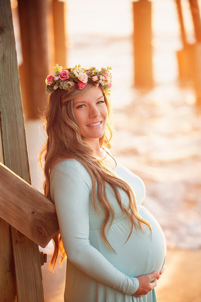 A pregnant woman in a flowing blue dress stands on a wooden pier, embracing the peaceful atmosphere of her surroundings.