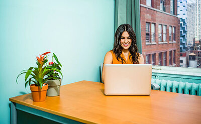 The image shows a smiling woman working on a laptop at a wooden desk. She has dark hair and is wearing a sleeveless orange top. To her left, there is a potted plant with green leaves and red accents, adding a pop of color to the scene. The room has a light blue wall and a large window through which daylight and a cityscape with buildings are visible. The woman appears to be engaged with her work, exuding a cheerful and productive atmosphere.