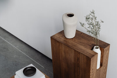 Modern minimalist wooden accent table with two white ceramic vases, one holding dried foliage, styled against a white wall on a polished concrete floor.
