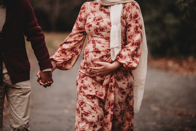 Celebrate the beauty of pregnancy with a stunning outdoor maternity photoshoot in Connecticut. This heartwarming image captures a glowing mother-to-be in a floral gown, holding hands with her partner on a scenic nature path. Book your elegant maternity session with Nicole Mele Photography to create timeless memories.