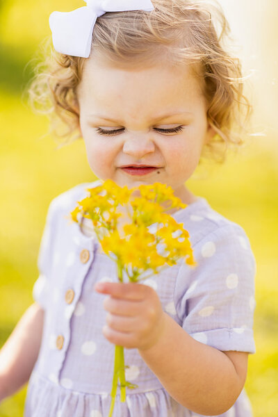 A little girl in a purple dress smells a yellow flower
